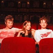 Three people sit in red theater seats, with the two on the sides leaning toward the person in the middle.